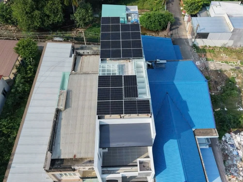 Toàn cảnh các cụm tấm pin trên mái nhà dài, xen giữa là khoảng mái và ô lấy sáng, bên cạnh những mái tôn xanh.